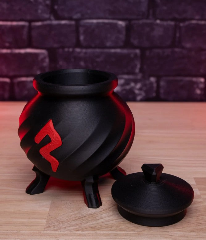 Black and red cauldron-shaped container with a lid on a wooden surface against a brick wall background
